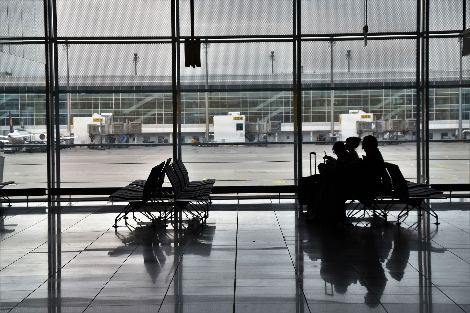 空港ロビーの窓際でスーツケースと共に搭乗を待つ旅行者のシルエット(イメージ)