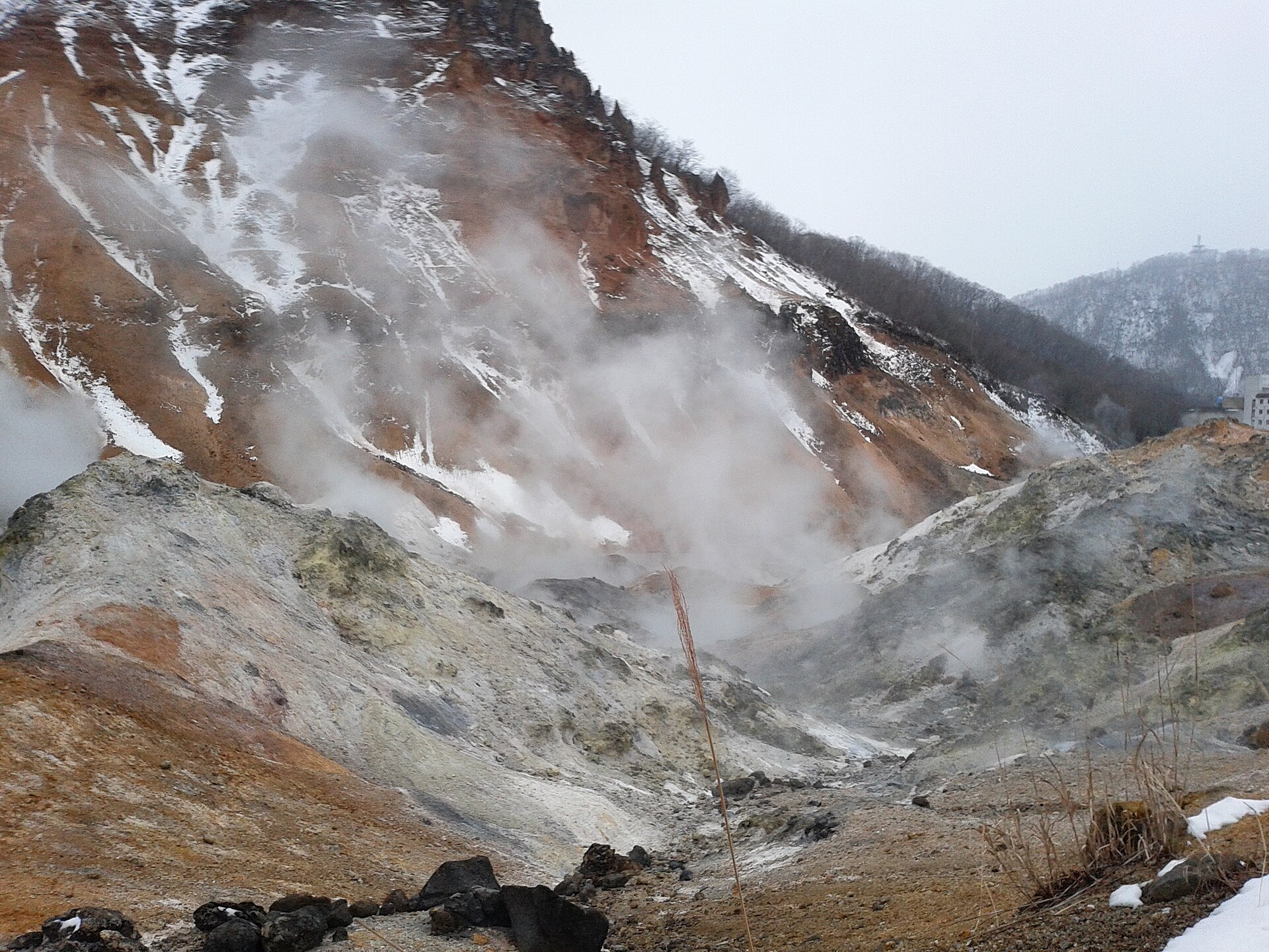 雪をかぶった赤茶色の山肌の前に白い噴気が立ちこめる登別地獄谷