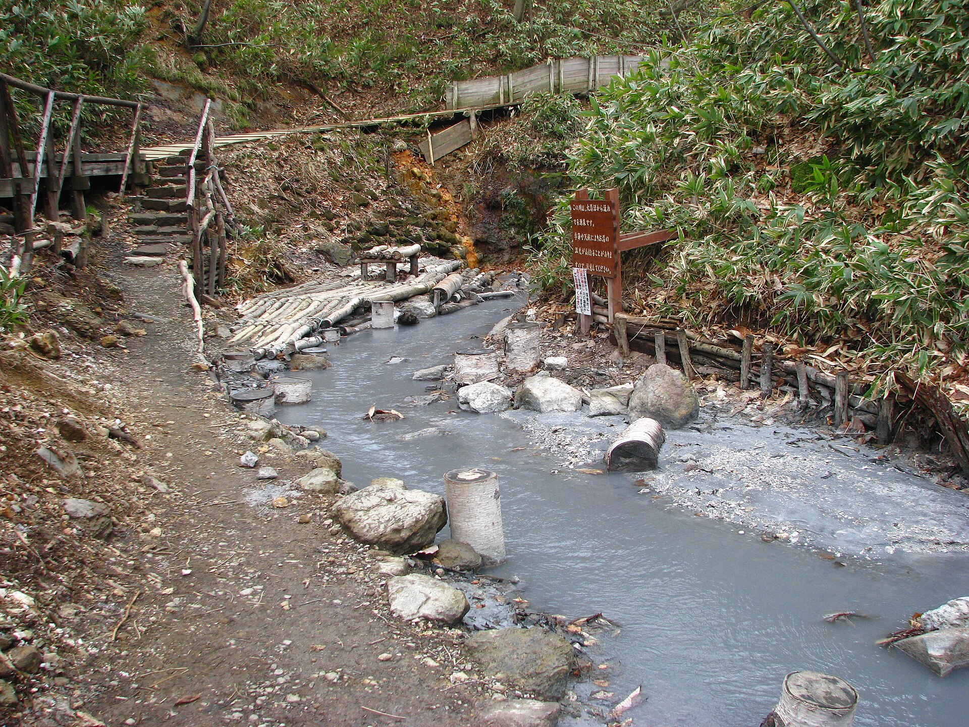 大湯沼川天然足湯。乳白色の温泉の川に丸太の腰掛けと木道の階段がある
