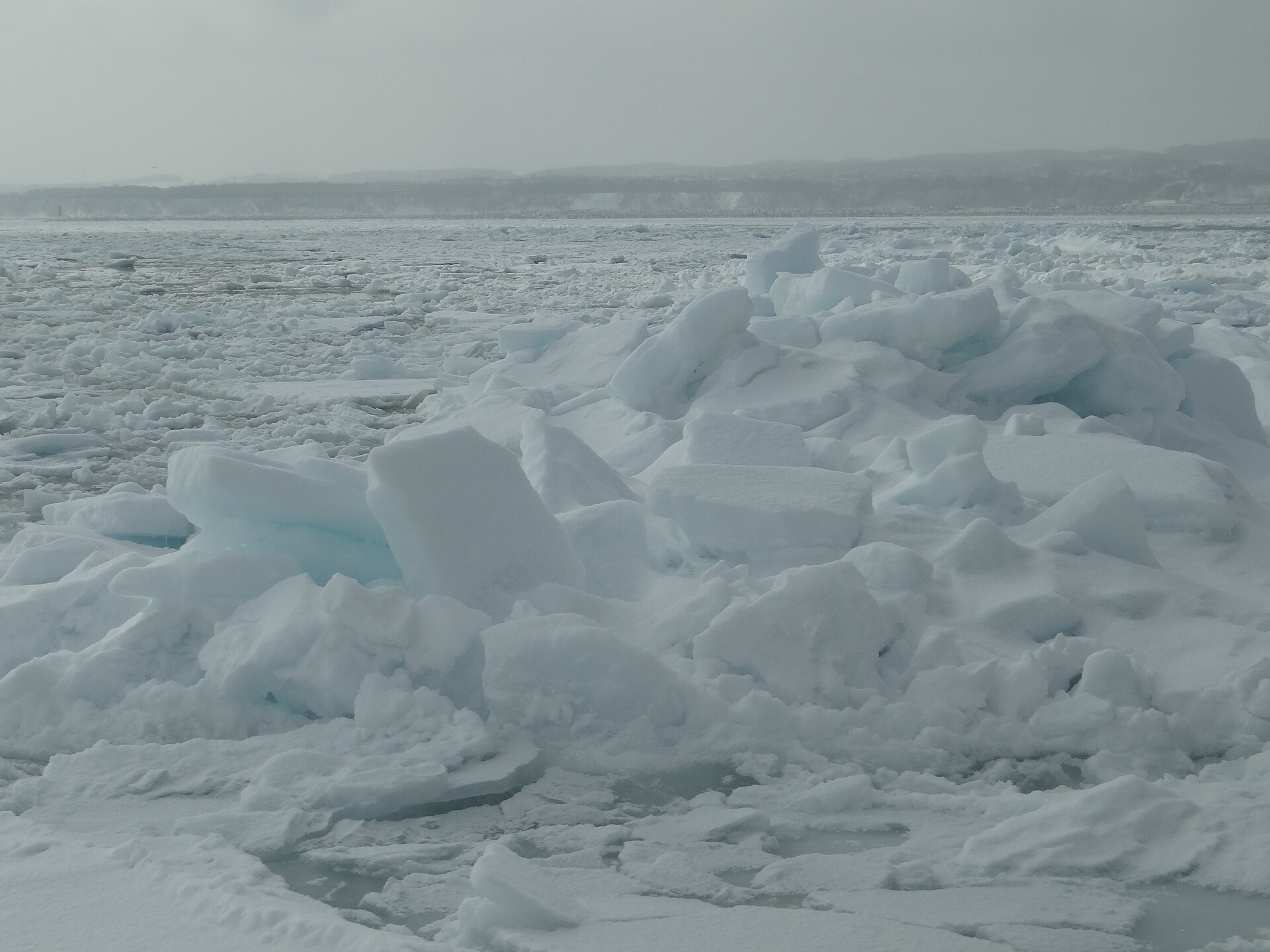 岸に押し寄せて積み重なった流氷の氷塊。割れた断面が青みを帯び、奥に網走の海岸が続く