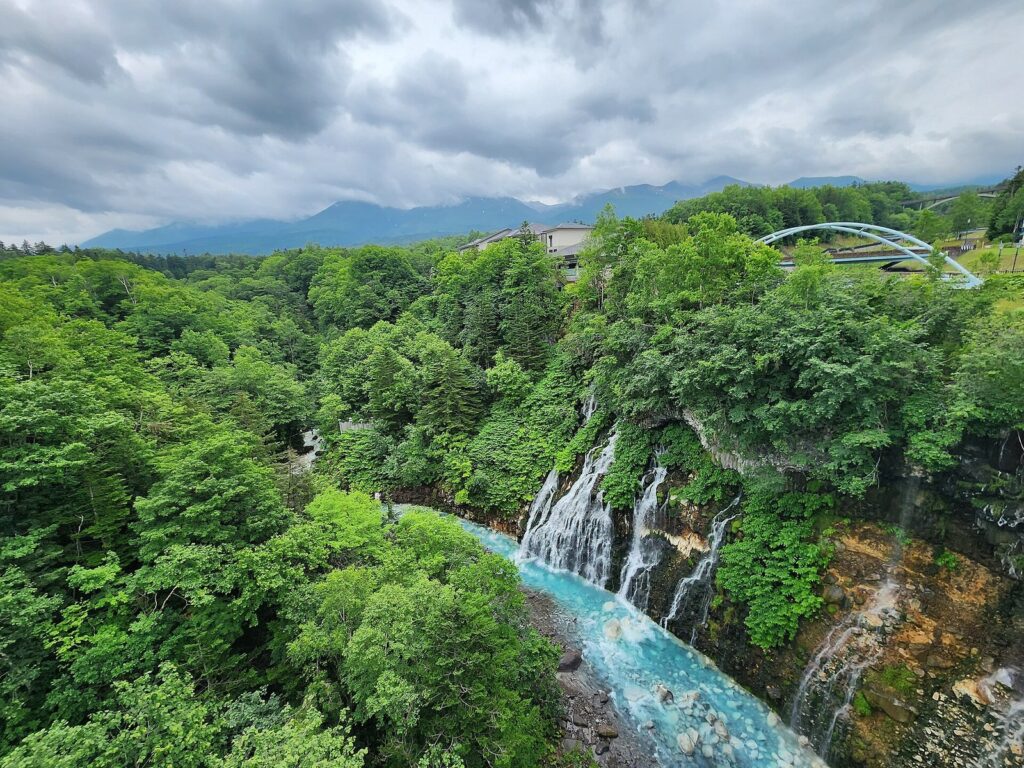 崖の岩肌から青い美瑛川へ流れ落ちる白ひげの滝と周囲の森