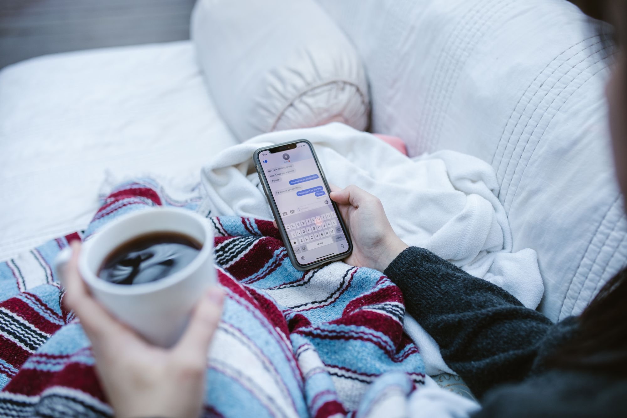 毛布にくるまりコーヒーを片手にスマートフォンを操作する人(イメージ)