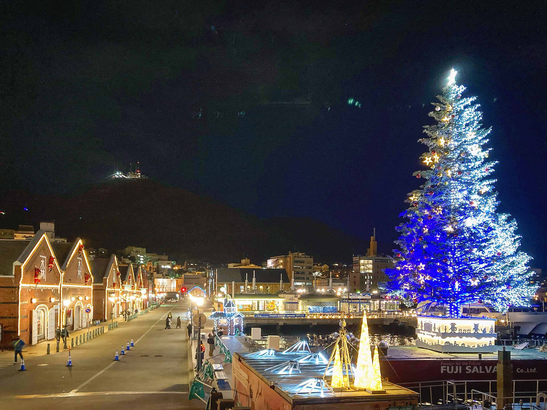 夜の金森赤レンガ倉庫群の前に浮かぶ青白く光る巨大クリスマスツリーと、奥に見える函館山