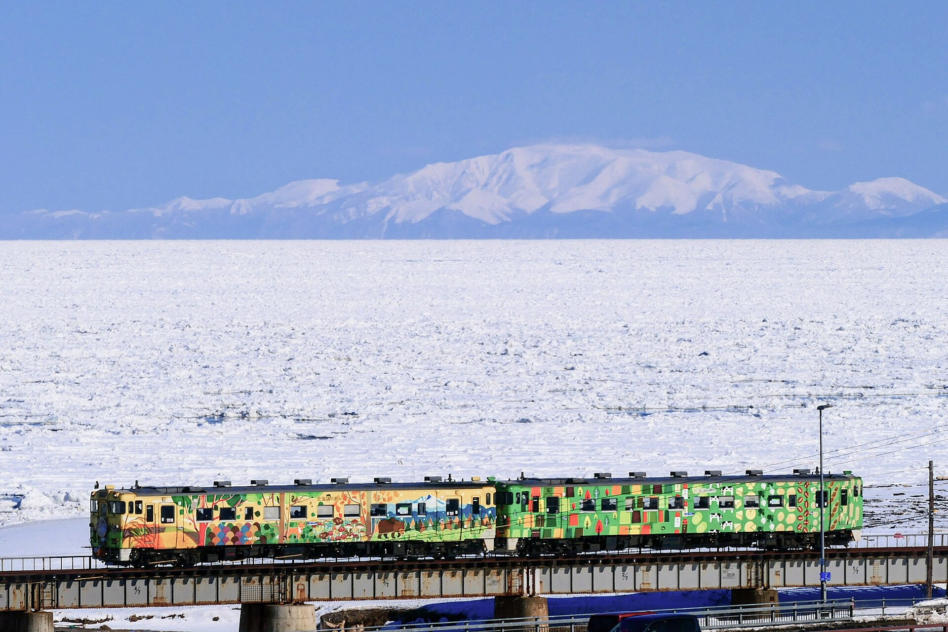 流氷で白く埋まった海と雪山を背景に、鉄橋を渡るカラフルな塗装の2両編成の列車