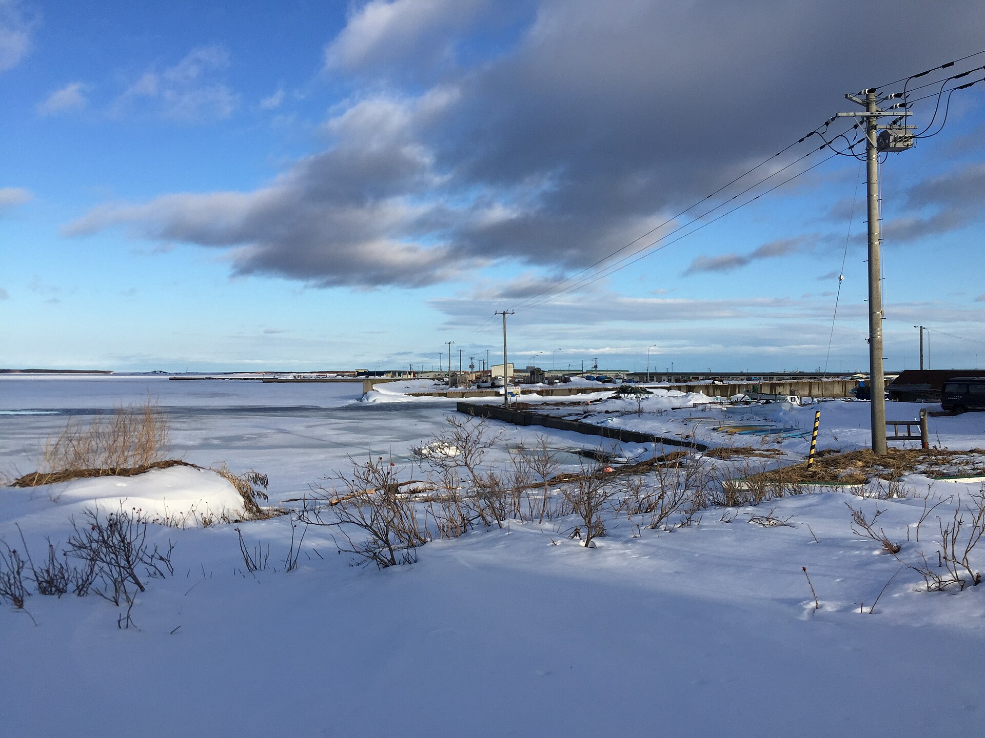 雪の積もった岸辺と氷の張った湖面。奥に小さな船着き場と電柱が見える冬の湖畔
