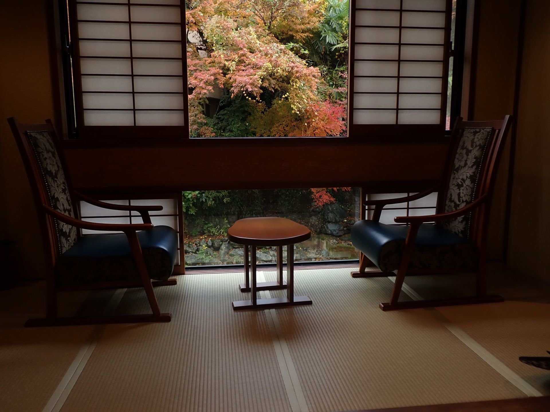 障子窓の向こうに紅葉の庭が見える、椅子とテーブルを置いた畳の部屋(イメージ)