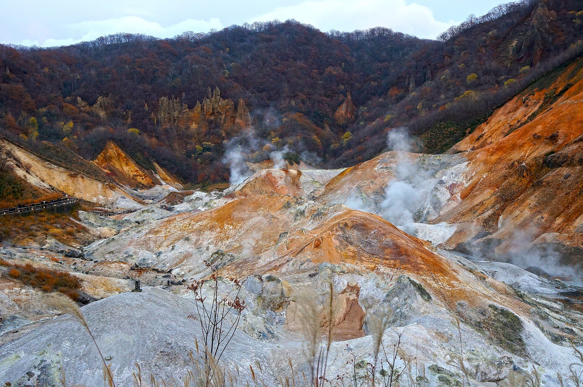 紅葉した山に囲まれ、黄褐色や白の岩肌から噴気が上がる秋の登別地獄谷